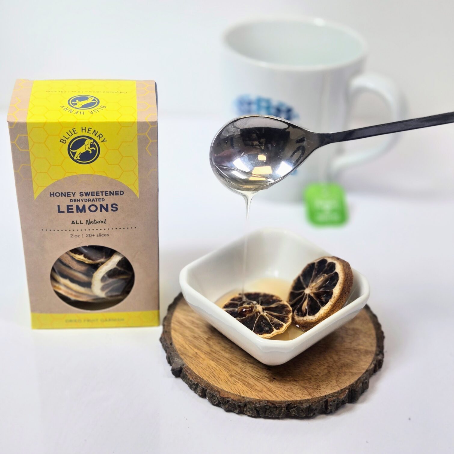Box of BLueHenry Honey Sweetened Lemons. A few slices are having a spoonful of honey poured over them with a cup of tea in the background